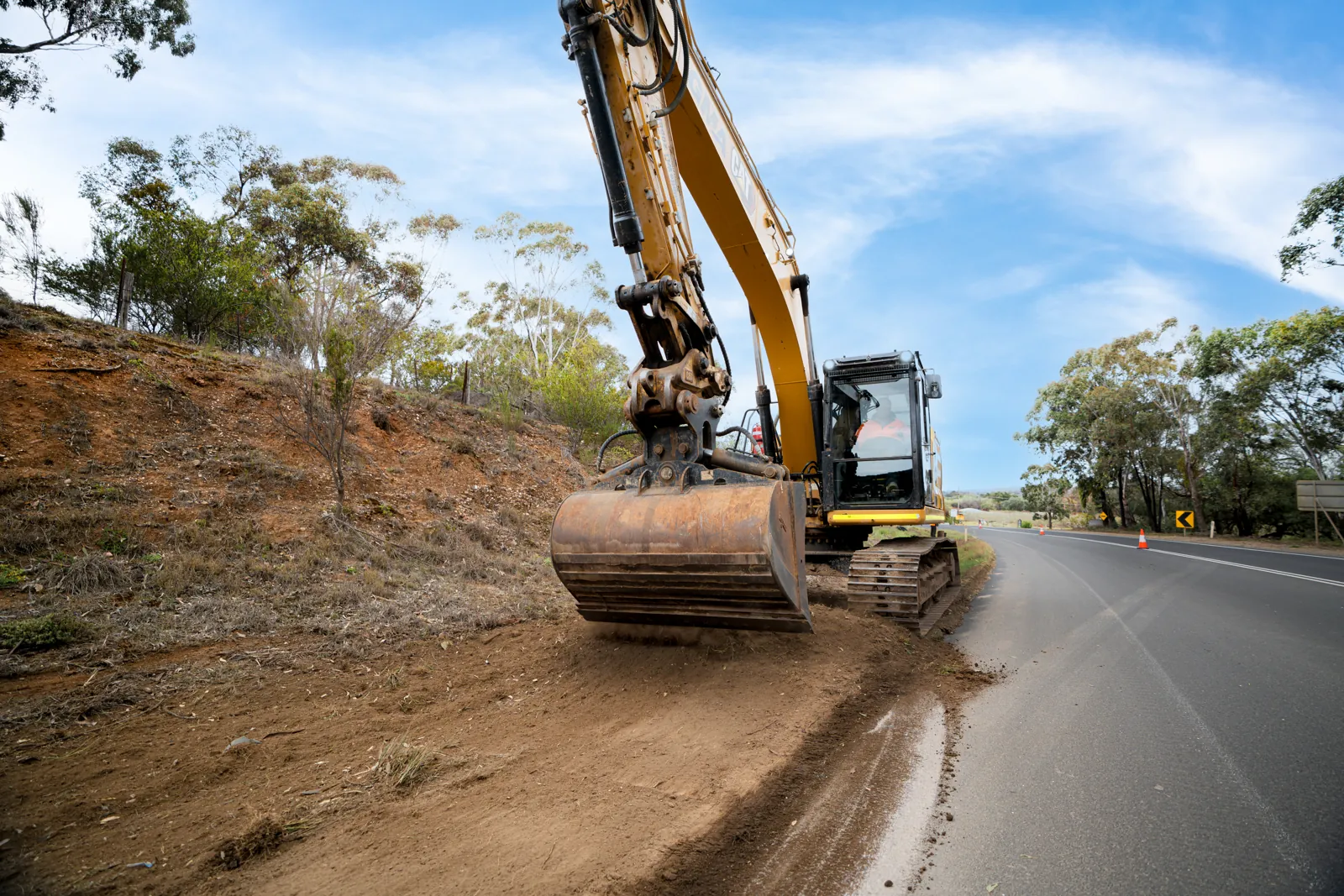 Excavator trimming a roadside batter beside a sealed road